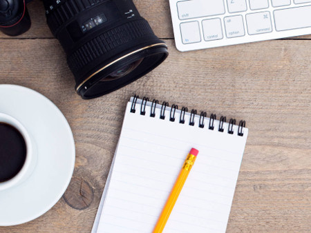 An image of a camera, keyboard, coffee and a notepad and pencil on a desk