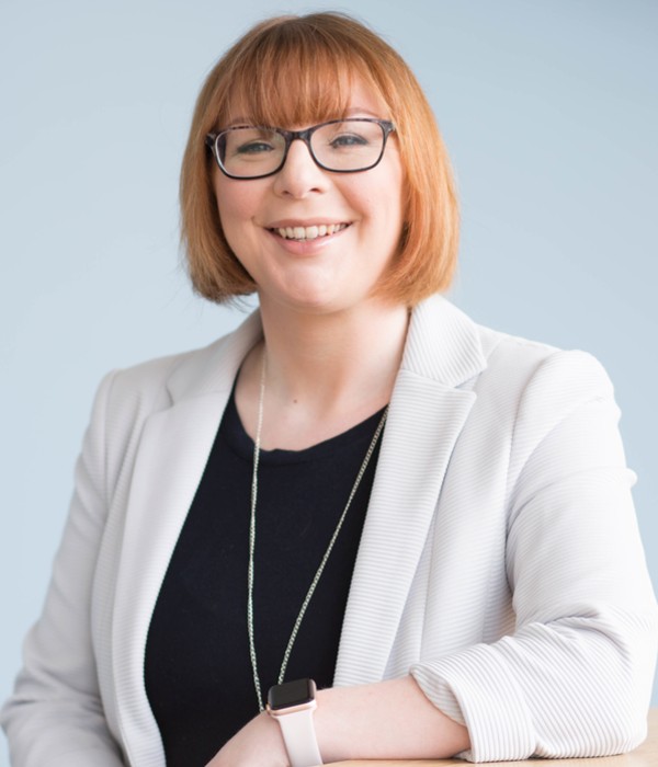 Dr Vikki McCall. Female with red bobbed hair cut, black glasses look straight to camera and wearing a white blazer and black top