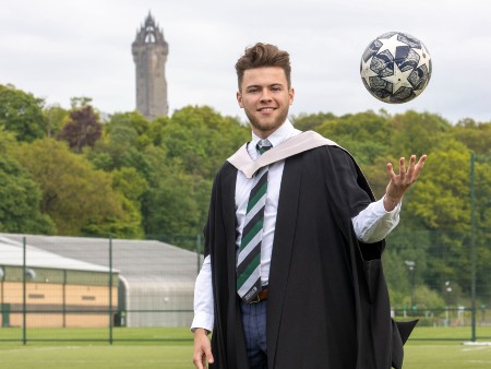 Footballer Craig Brown tosses a football with one hand while looking to camera, wearing his graduation robes with the Wallace Monument seen in the background