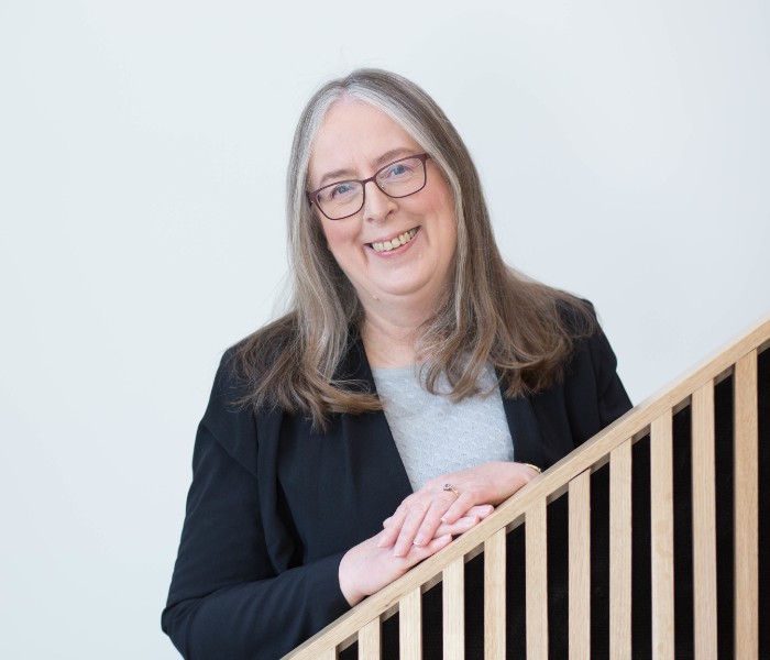 Prof Alison Bowes has shoulder length grey hair and wears dark, square glasses. She is looking to the camera smiling with her hands clasped leaning on the bannister of the staircase she is standing on