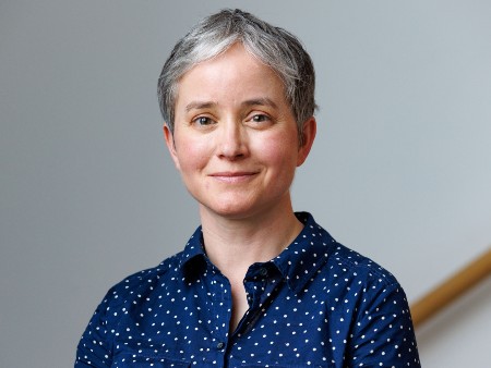 Portrait photograph of Kirstie Blair. She has short grey hair and is wearing a blue polka dot blouse. She is pictured indoors, against a grey background.