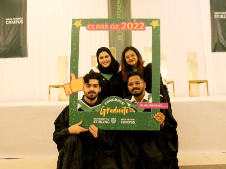 A group of four graduates from Stirling's RAK campus pose using a prop - an oversized green photo frame