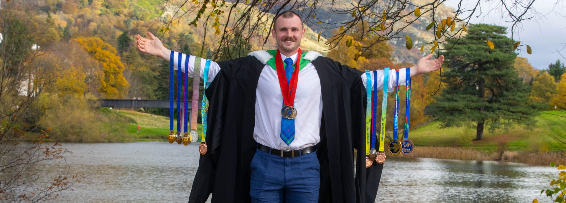 Ross poses next to loch in graduation gown holding medals