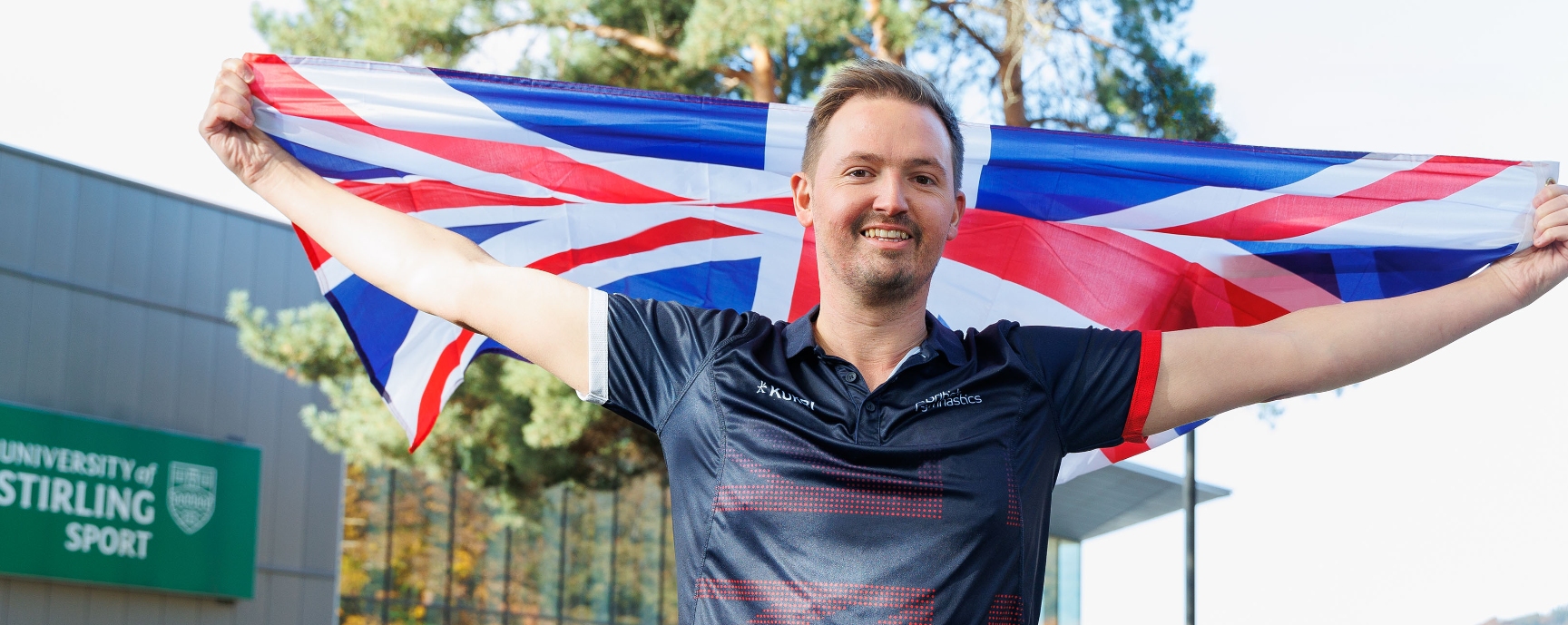 Paul Greaves holds a Great Britain flag outstretched above his head outside the University's Sports Centre